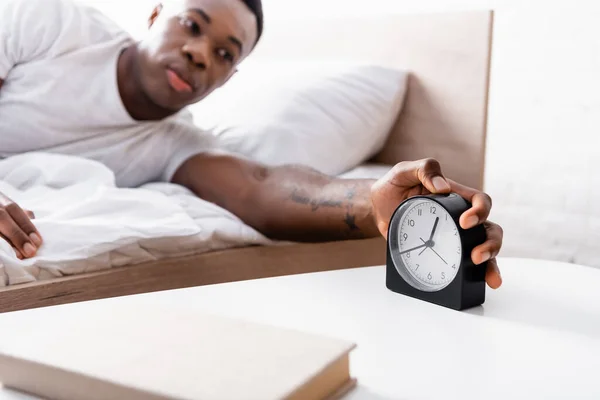 Clock in hand of african american man on bed on blurred background — Stock Photo