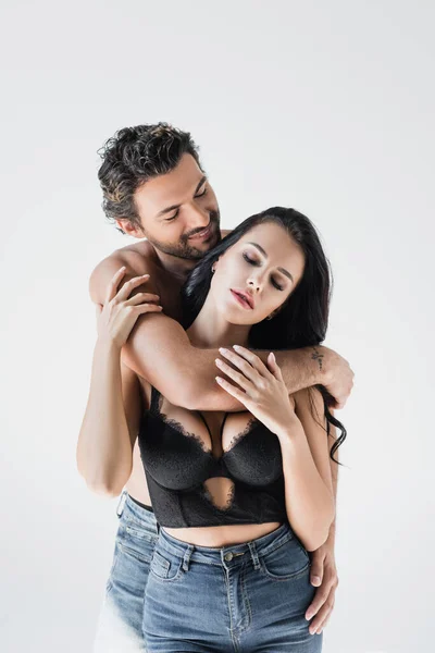 Lächelnder Mann umarmt brünette Freundin in BH und Jeans isoliert auf grau — Stockfoto