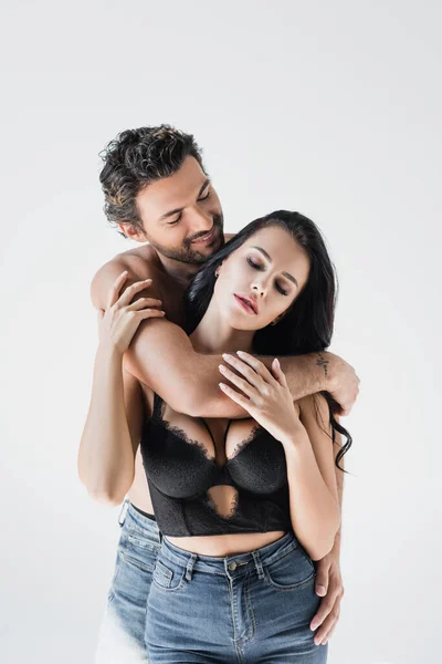 Uomo sorridente che abbraccia la ragazza bruna in reggiseno e jeans isolati sul grigio — Foto stock