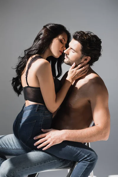 Sexy woman kissing muscular boyfriend in jeans on chair on grey background — Stock Photo