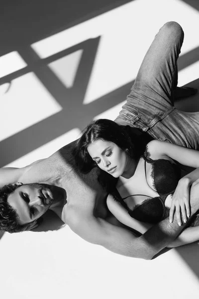 Monochrome shot of shirtless man hugging sexy woman while lying on white background with shadow — Stock Photo