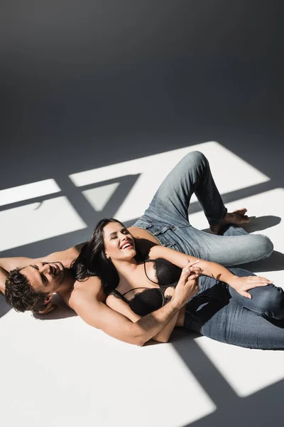 Cheerful woman in bra holding hand of shirtless boyfriend in jeans on grey background with shadow — Stock Photo