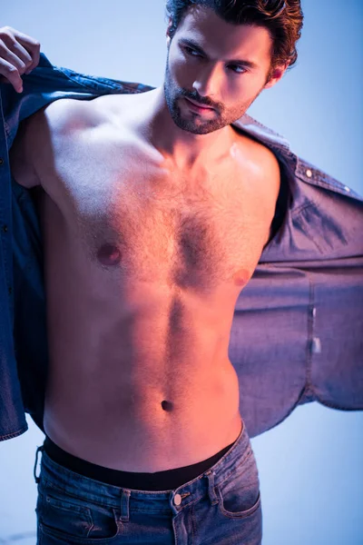 Young man taking off denim shirt on blue background — Stock Photo