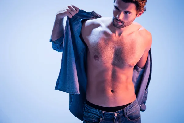 Muscular man taking off denim shirt and looking away on blue background — Stock Photo