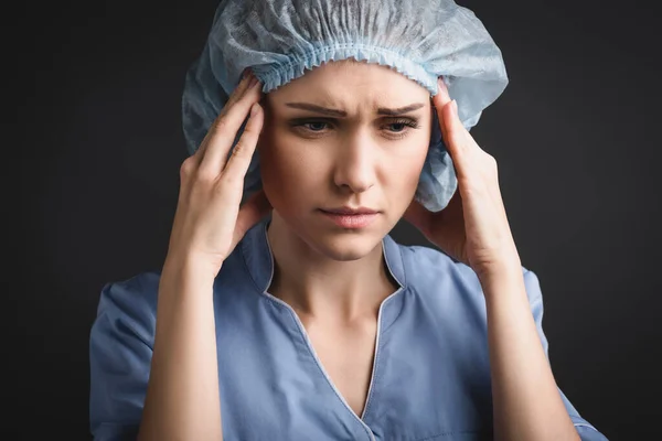 Enfermera cansada en gorra médica que sufre de dolor de cabeza aislado en gris oscuro - foto de stock