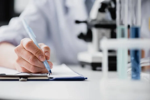 Vista parcial de la escritura de la enfermera en el portapapeles cerca del microscopio sobre fondo borroso - foto de stock