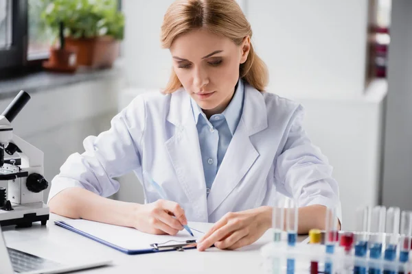 Scientist writing on clipboard near microscope and test tubes on blurred background — Stock Photo