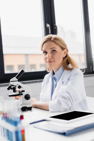 Científico feliz en la bata blanca mirando la cámara cerca del microscopio y la tableta digital con la pantalla en blanco en el escritorio - foto de stock