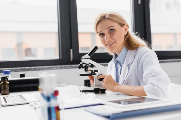 Веселый ученый в белом халате смотрит в камеру рядом с микроскопом и цифровым планшетом на столе — стоковое фото