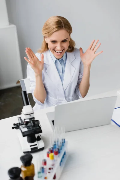 Cheerful scientist in white coat looking at laptop near microscope on desk — Stock Photo