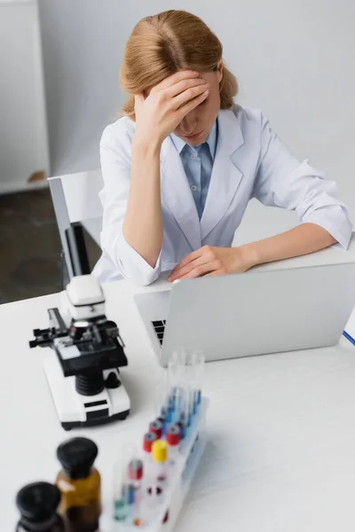 Sad scientist  in white coat covering face near laptop and microscope on desk — Stock Photo