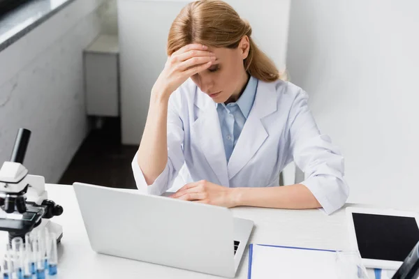 Upset scientist in white coat covering face near laptop and microscope on desk — Stock Photo