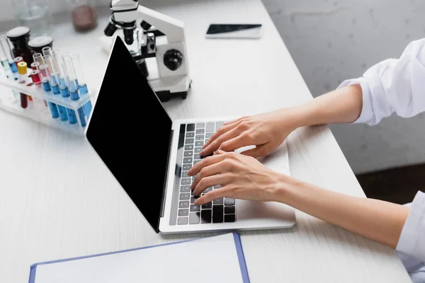 Vista recortada del científico escribiendo en el ordenador portátil con pantalla en blanco cerca del microscopio en el escritorio - foto de stock