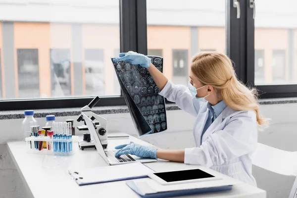 Scientist in medical mask looking at x-ray near devices on desk — Stock Photo