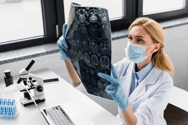 Wissenschaftler in medizinischer Maske betrachtet röntgennahe Geräte und Mikroskop auf dem Schreibtisch — Stockfoto
