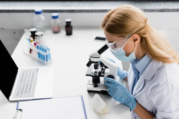 Cientista em máscara médica olhando através de microscópio na mesa — Fotografia de Stock