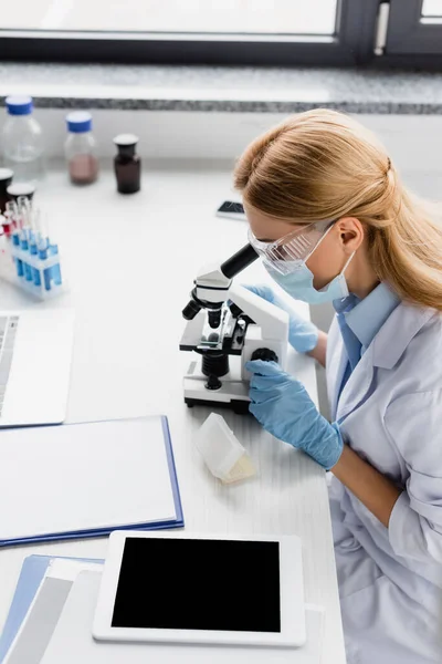 Scientist in medical mask and goggles looking through microscope on desk — Stock Photo