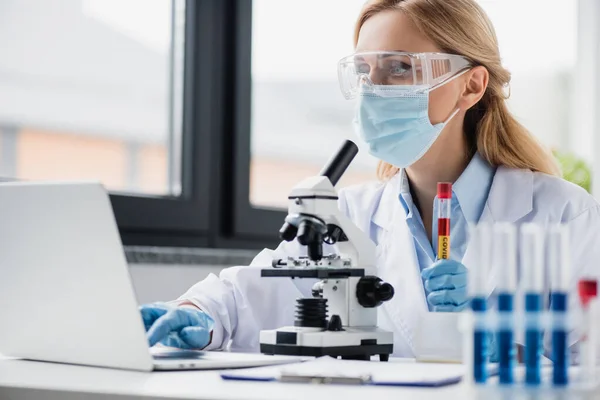 Cientista em máscara médica e óculos segurando tubo de ensaio com letras vívidas e usando laptop — Fotografia de Stock