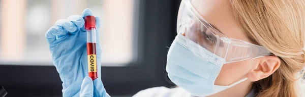 Científico en máscara médica y gafas mirando tubo de ensayo con letras covid, pancarta - foto de stock