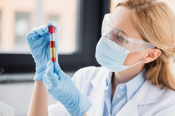 Científico en máscara médica y gafas mirando el tubo de ensayo con letras covid en el laboratorio - foto de stock