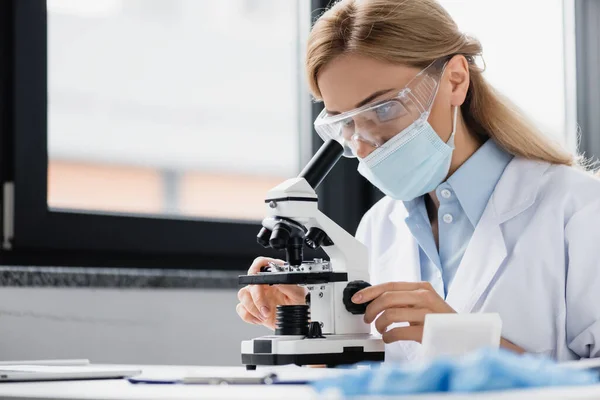 Científico en máscara médica y gafas mirando a través del microscopio - foto de stock