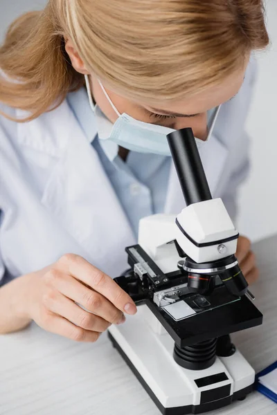 Female scientist in medical mask looking through microscope — Stock Photo