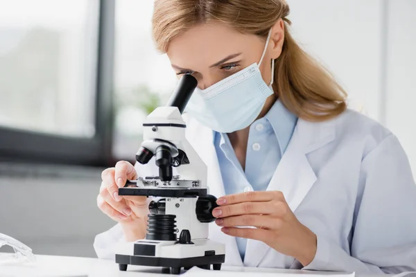 Especialista en mascarilla médica mirando a través del microscopio en laboratorio - foto de stock