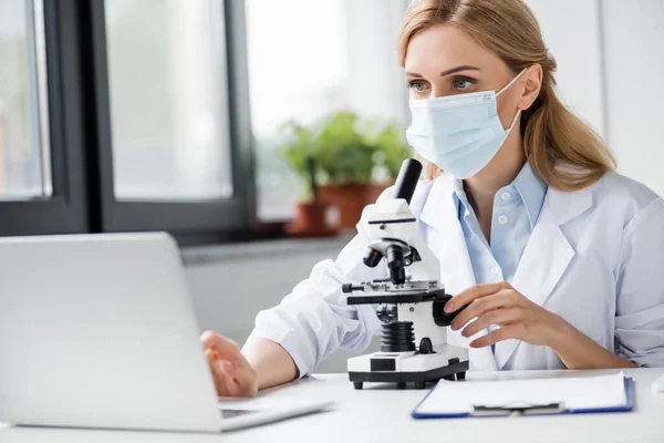 Especialista em máscara médica usando laptop perto de microscópio na mesa — Fotografia de Stock