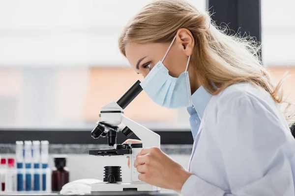 Especialista en mascarilla médica mirando a través del microscopio en laboratorio - foto de stock