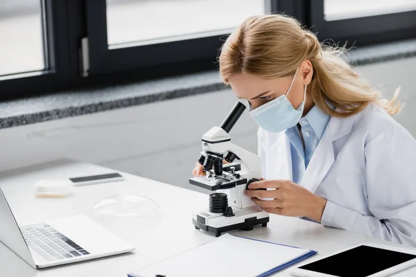 Scientifique en masque médical regardant à travers le microscope près de gadgets avec écran blanc sur le bureau — Photo de stock