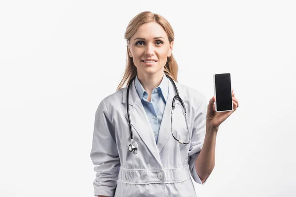 Fröhliche Krankenschwester in weißem Mantel lächelt, während sie Smartphone mit leerem Bildschirm auf weiß isoliert hält — Stockfoto