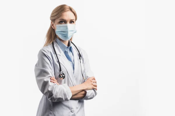 Enfermera en máscara médica y bata blanca de pie con brazos cruzados aislados en blanco - foto de stock