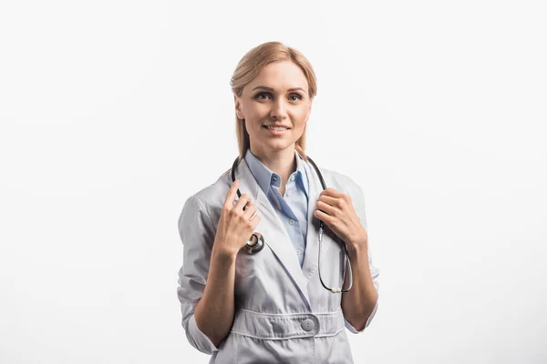 Enfermeira alegre em casaco branco ajustando estetoscópio isolado no branco — Fotografia de Stock