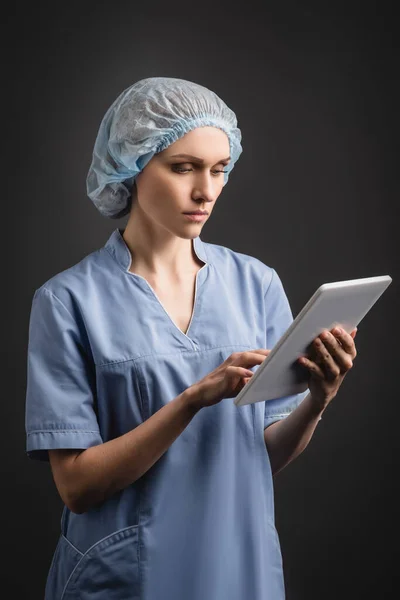 Nurse in medical cap and uniform using digital tablet isolated on dark grey — Stock Photo
