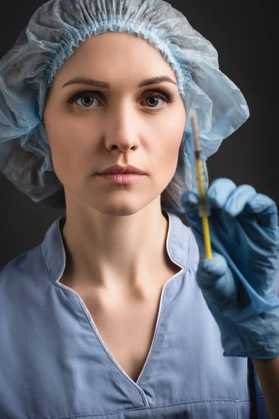 Enfermera con gorra médica y jeringa uniforme con vacuna en primer plano borrosa aislada en gris oscuro - foto de stock