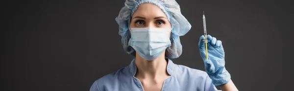 Медсестра в медицинской маске держит шприц с вакциной, изолированной на темно-сером, баннер — стоковое фото