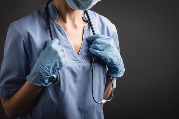 Visão recortada do enfermeiro em máscara médica e luvas de látex ajustando estetoscópio isolado em cinza escuro — Fotografia de Stock