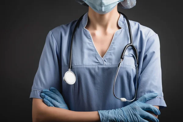 Vista recortada de la enfermera en máscara médica y guantes de látex de pie con brazos cruzados aislados en gris oscuro - foto de stock