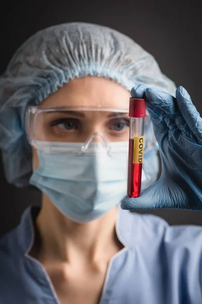 Пробирка с ковидовой надписью на руке медсестры в медицинском колпаке, маске и очках на размытом фоне, изолированном на темно-сером — стоковое фото