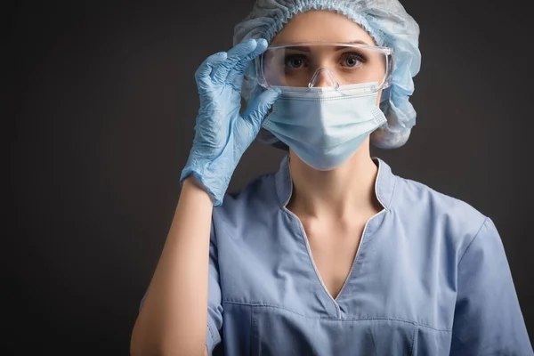 Enfermeira em tampa médica e máscara ajustando óculos enquanto olha para a câmera isolada em cinza escuro — Fotografia de Stock
