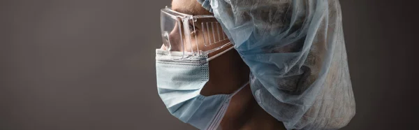 Side view of nurse in medical cap, mask and goggles looking away isolated on dark grey, banner — Stock Photo