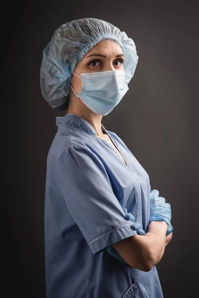 Enfermera en gorra médica y máscara de pie con los brazos cruzados y mirando a la cámara aislada en gris oscuro - foto de stock