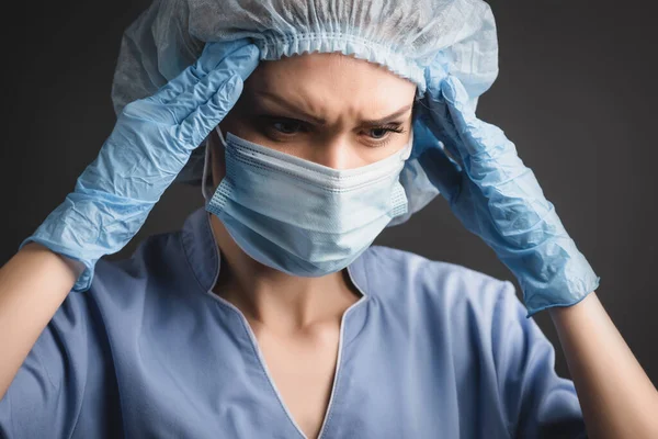 Enfermeira em tampa médica e máscara que sofre de enxaqueca isolada em cinza escuro — Fotografia de Stock