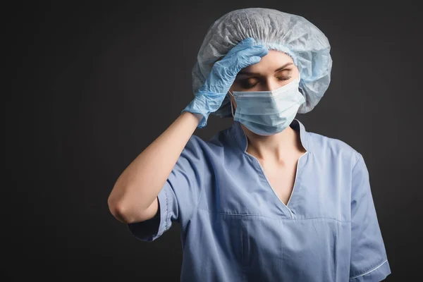 Tired nurse in medical cap and mask suffering from headache isolated on dark grey — Stock Photo