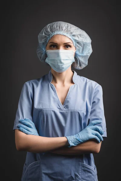 Nurse in latex gloves, medical mask and cap standing with crossed arms isolated on dark grey — Stock Photo