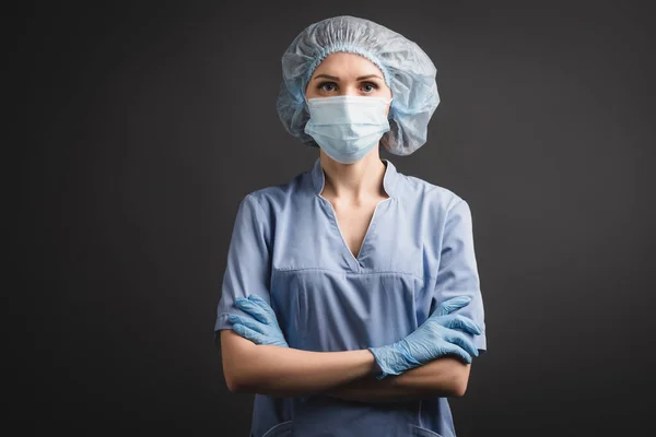 Enfermera en guantes de látex, gorra médica y máscara de pie con brazos cruzados aislados en gris oscuro - foto de stock