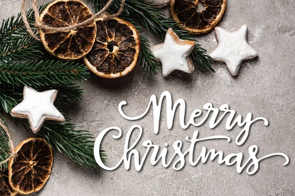 Top view of pine branch with dried orange pieces and cookies near merry christmas lettering on grey background — Stock Photo