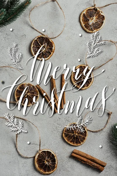 Top view of dried orange pieces with strings, cinnamon sticks and pine branches near merry christmas lettering — Stock Photo