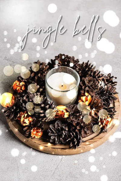 Ароматизированная свеча с сосновыми шишками на деревянной тарелке рядом с колокольчиками, надпись на сером фоне — стоковое фото