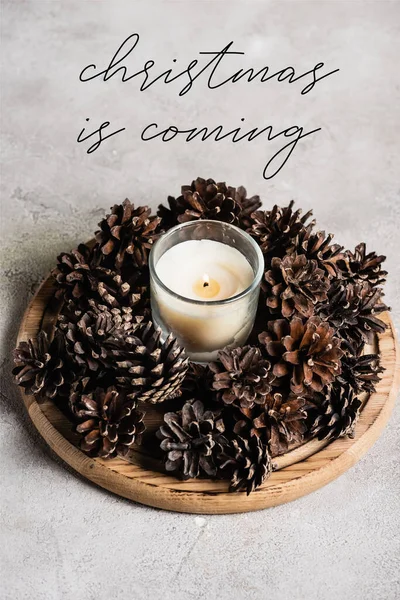 Vela perfumada con conos de pino en placa de madera cerca de Navidad en letras que vienen sobre fondo gris - foto de stock